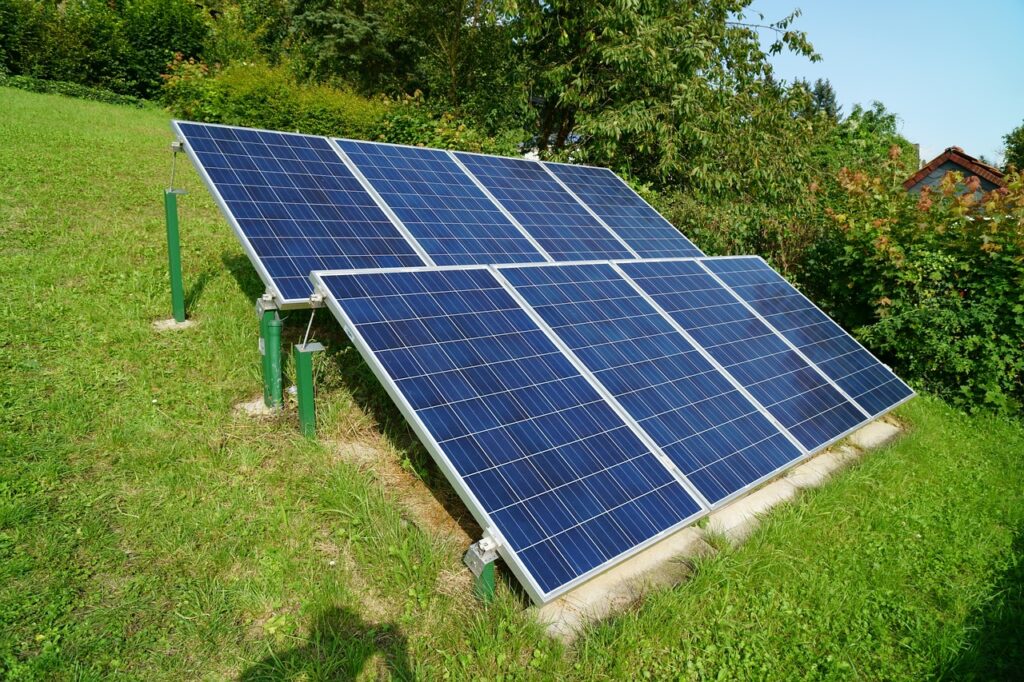 Freistehende Photovoltaikanlage im Garten mit mehreren blauen Solarmodulen, die auf einer geneigten Metallkonstruktion montiert sind. Die Anlage steht auf einer grünen Wiese, umgeben von Bäumen und Sträuchern. Im Hintergrund ist ein Haus mit rotem Dach teilweise sichtbar. Der Himmel ist klar und sonnig.