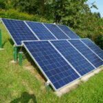 Freistehende Photovoltaikanlage im Garten mit mehreren blauen Solarmodulen, die auf einer geneigten Metallkonstruktion montiert sind. Die Anlage steht auf einer grünen Wiese, umgeben von Bäumen und Sträuchern. Im Hintergrund ist ein Haus mit rotem Dach teilweise sichtbar. Der Himmel ist klar und sonnig.